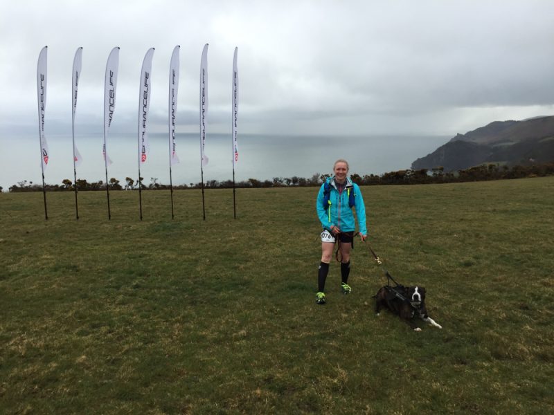 A happy Canicrosser and her dog standing on the top of a cliff. It is cloudy and grey